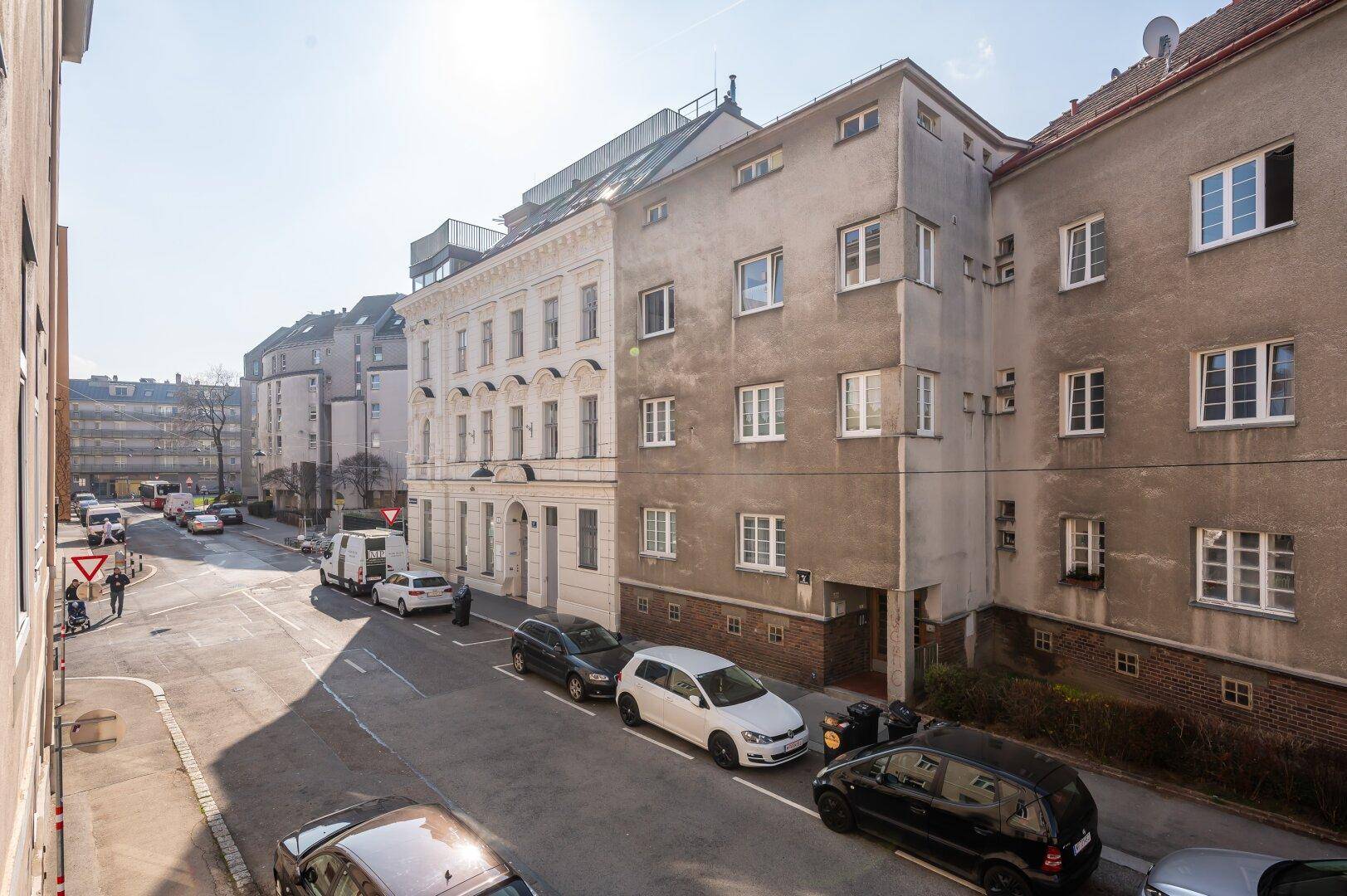 Sonnenbeschienene Straße in einem städtischen Viertel mit geparkten Autos und Wohngebäuden.