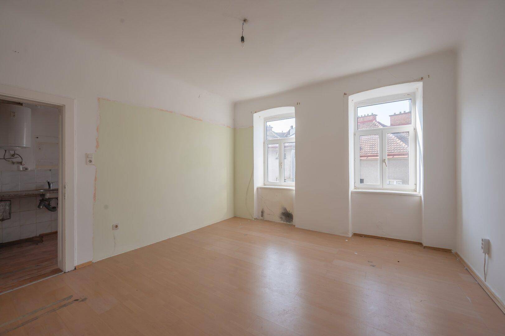 Leeres Zimmer mit Holzboden und zwei Fenstern, das Renovierungsbedarf aufweist.
