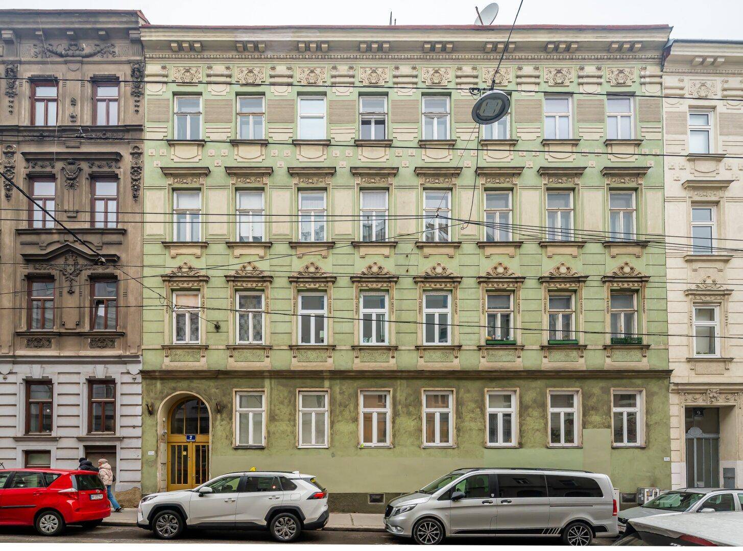 Historische Fassade eines Altbaus mit dekorativen Elementen und parkenden Autos auf der Straße.