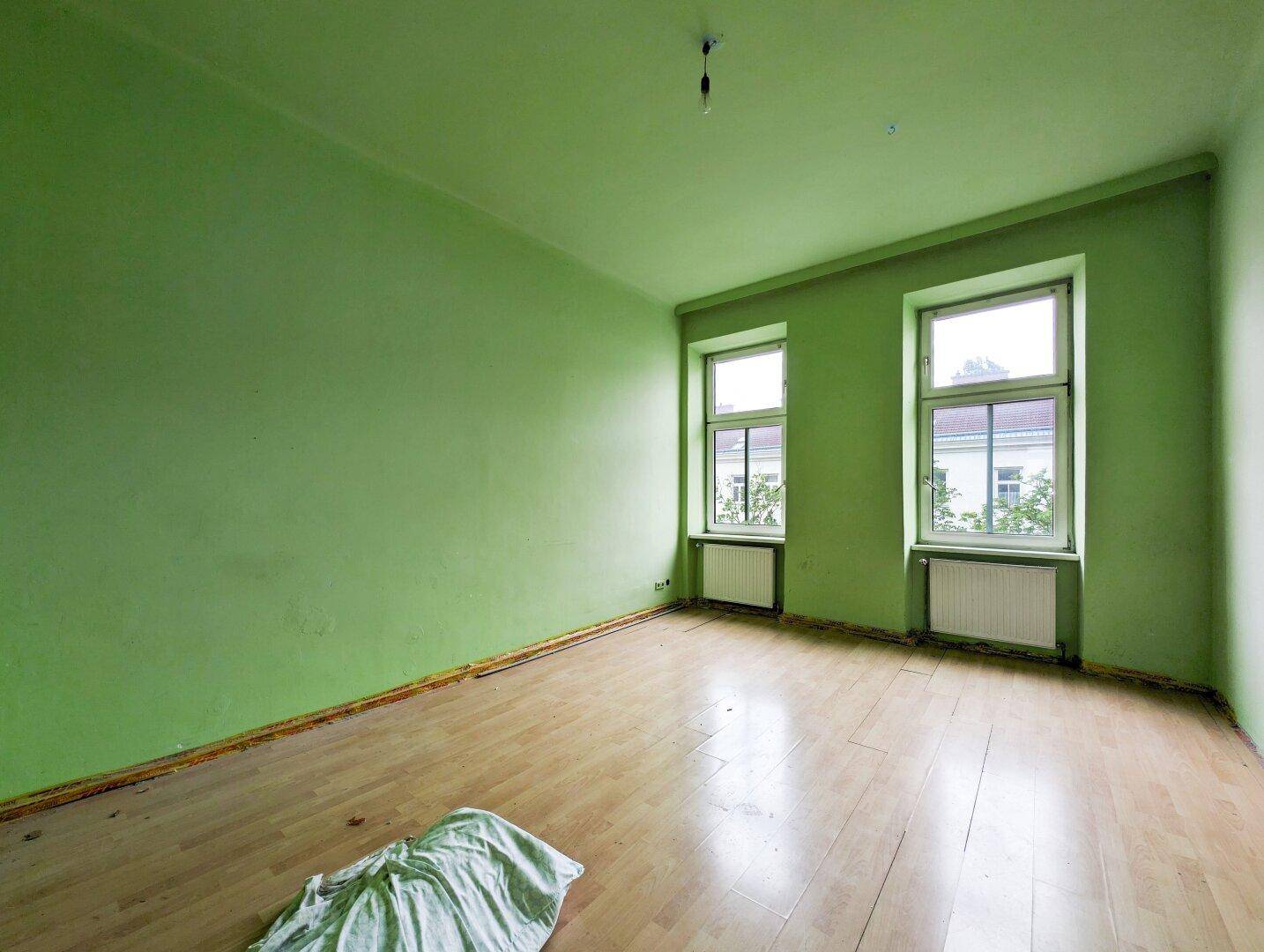 Leeres Zimmer mit hellgrünen Wänden, Holzboden und zwei Fenstern für natürliches Licht.