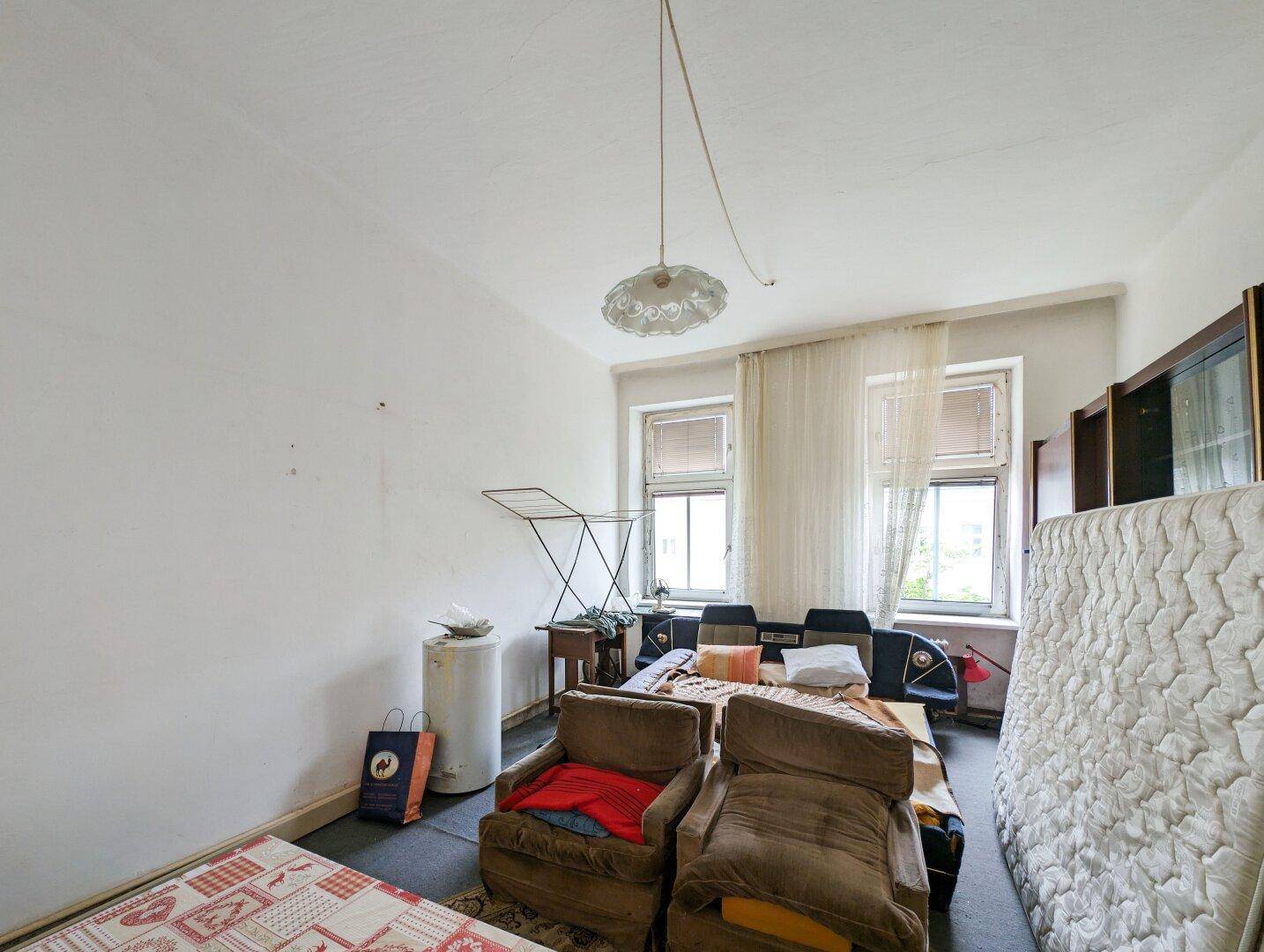 Unaufgeräumtes Wohnzimmer mit alten Möbeln, Matratze und Wäscheständer, in ursprünglichem Zustand.
