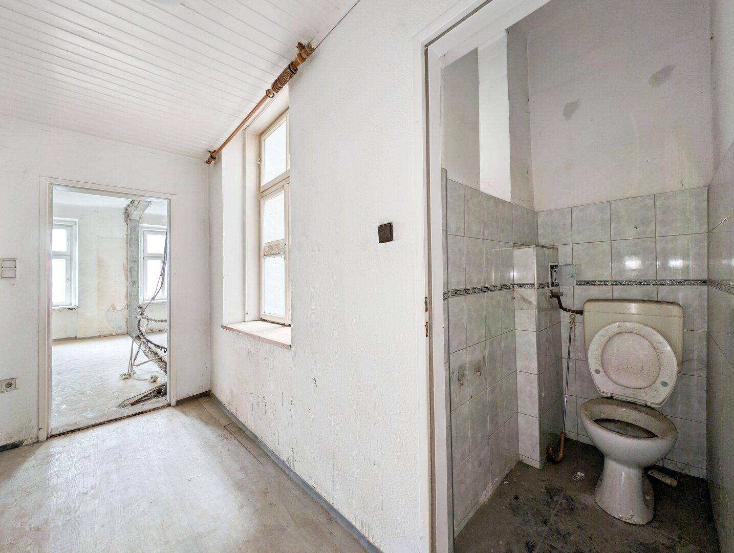 Kleiner WC-Bereich mit einfacher Toilette und Fliesen, angrenzend an einen weiteren Raum.