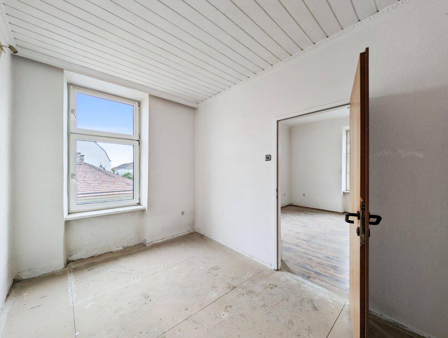 Helles Zimmer mit Holzdecke und einem Fenster, das einen Blick auf die Dächer bietet.