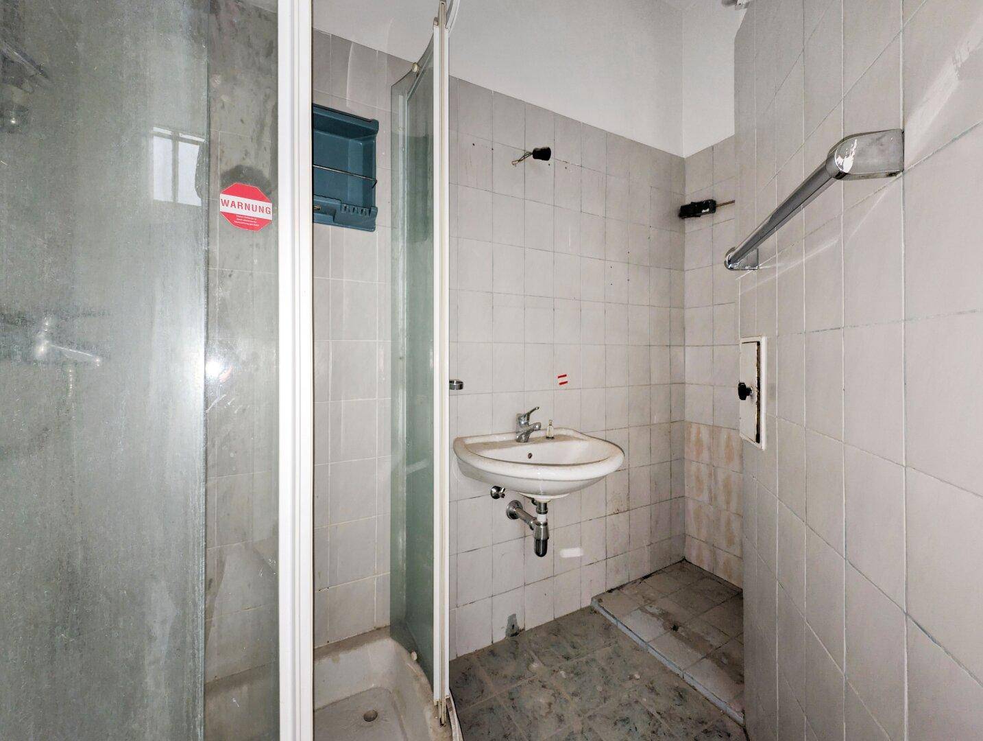 Einfaches Badezimmer mit Duschkabine, Waschbecken und weißen Fliesen, renovierungsbedürftig.