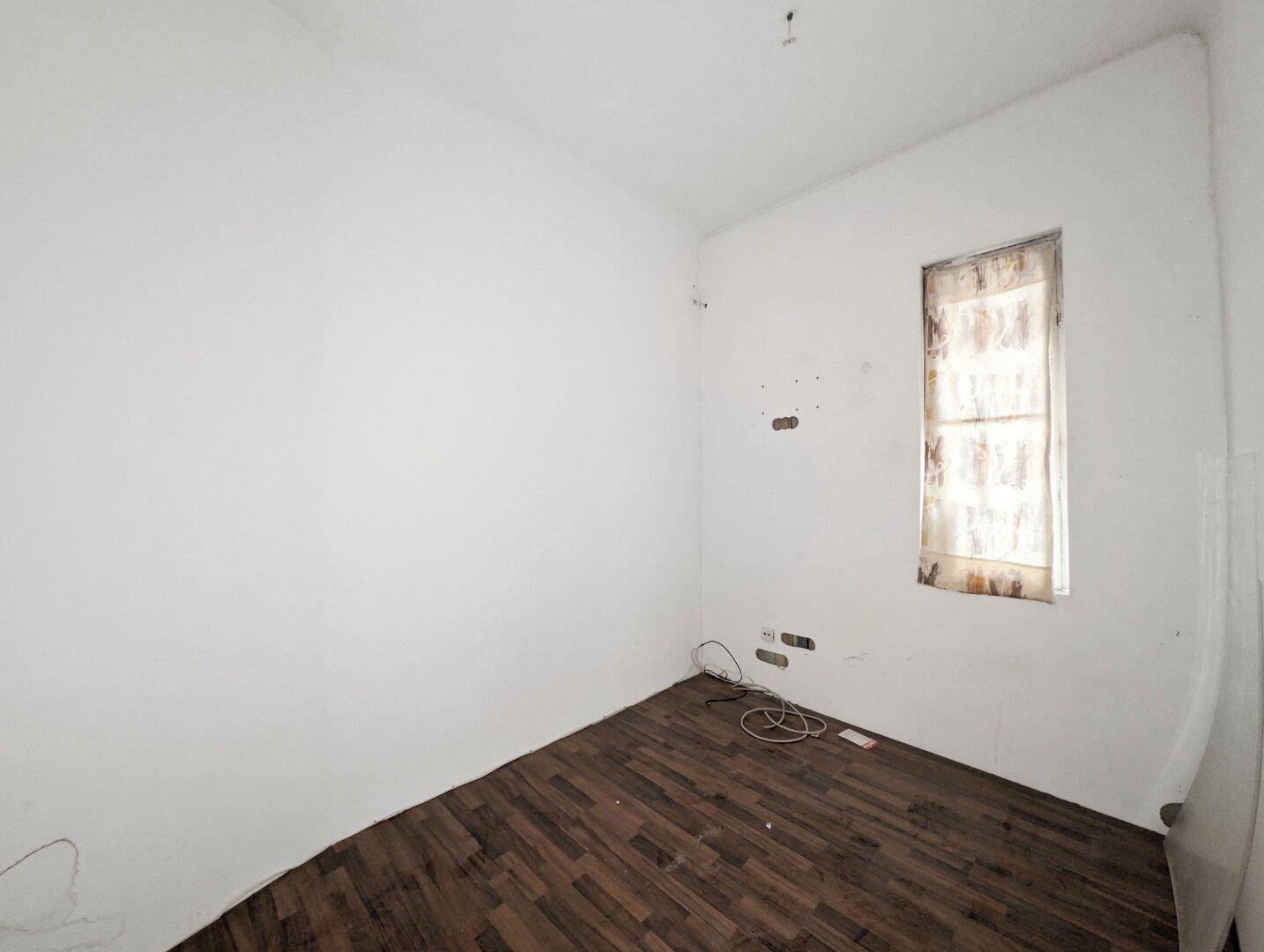 Kleines Zimmer mit dunklem Laminatboden und einem Fenster, ideal als Schlaf- oder Arbeitszimmer.