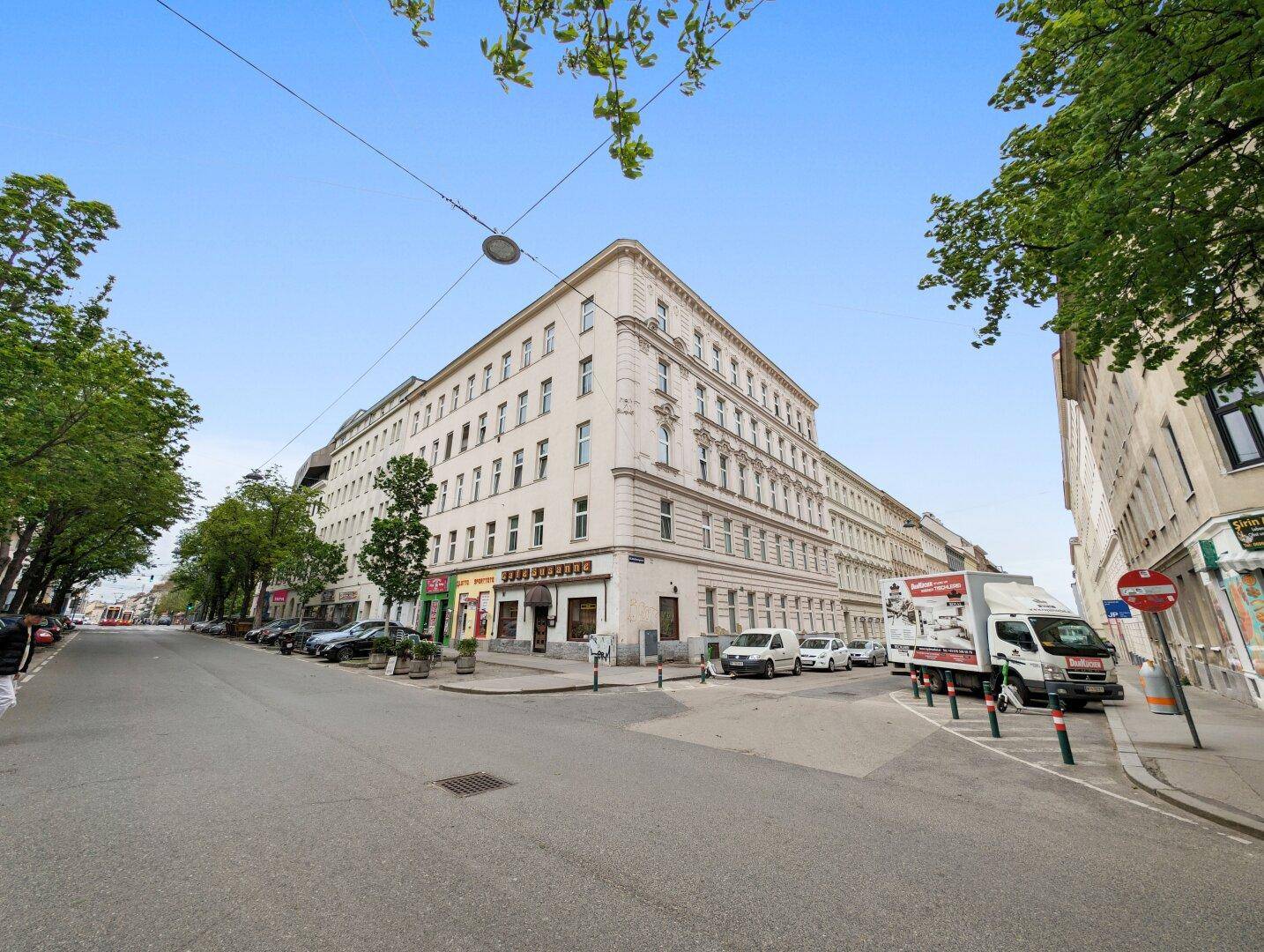 Historisches Eckgebäude in urbaner Umgebung mit Geschäften und parkenden Autos an der Straße.