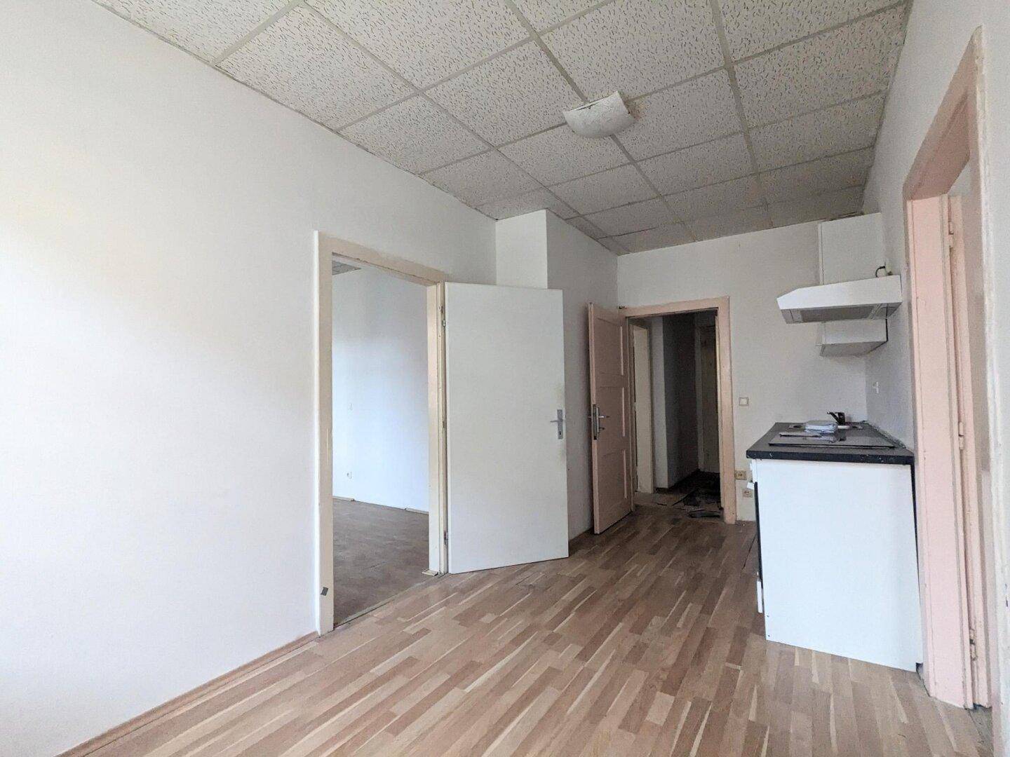 Einfache Küche mit Holzfußboden, kleiner Küchenzeile und Zugang zu weiteren Räumen.