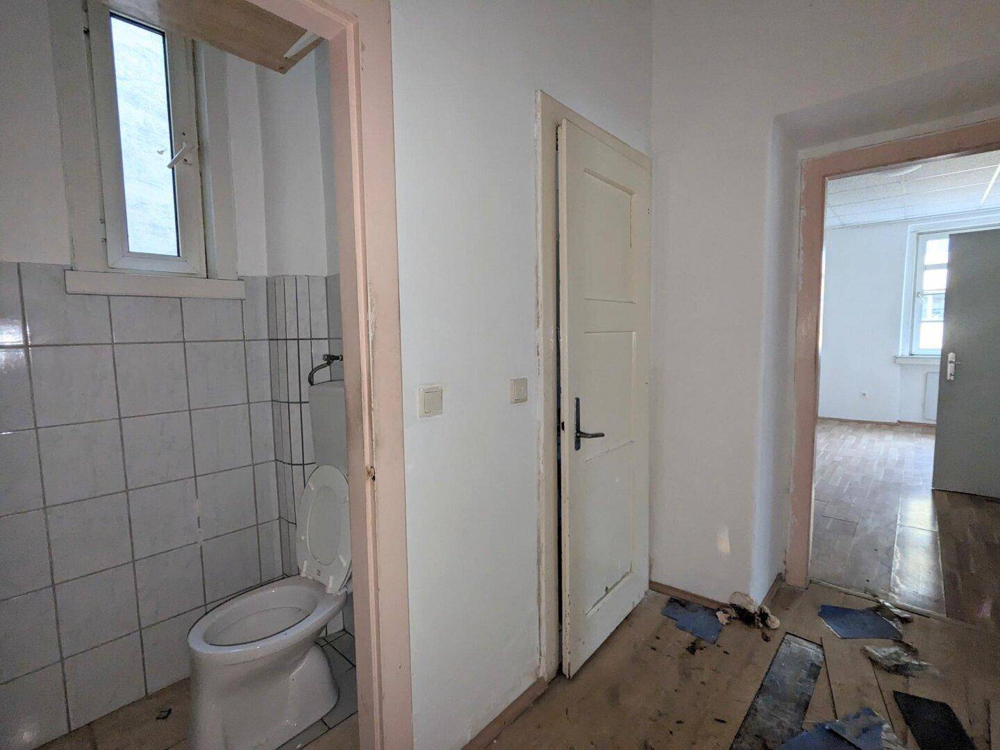 Flur führt zu einem WC mit weißen Fliesen, Fenster und sichtbaren Schäden am Boden.