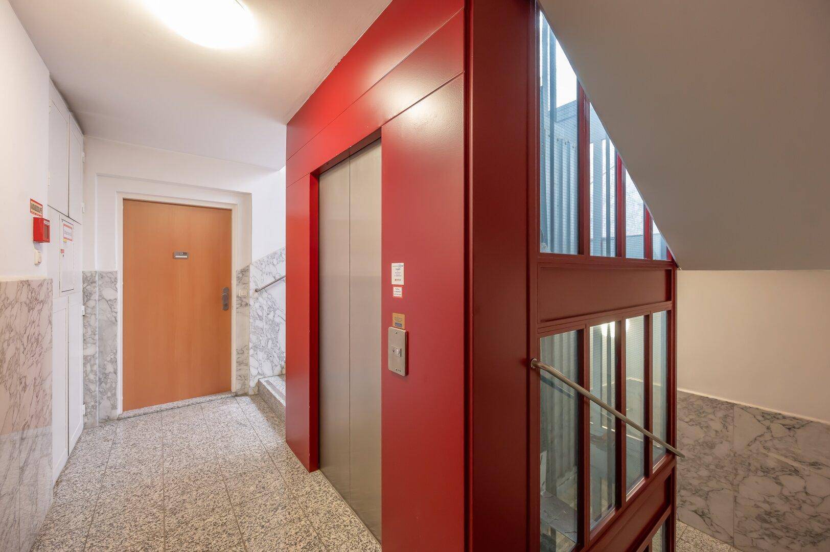 Moderner Eingangsbereich mit Aufzug und Treppenhaus, gestaltet mit roten Akzenten und Marmor.