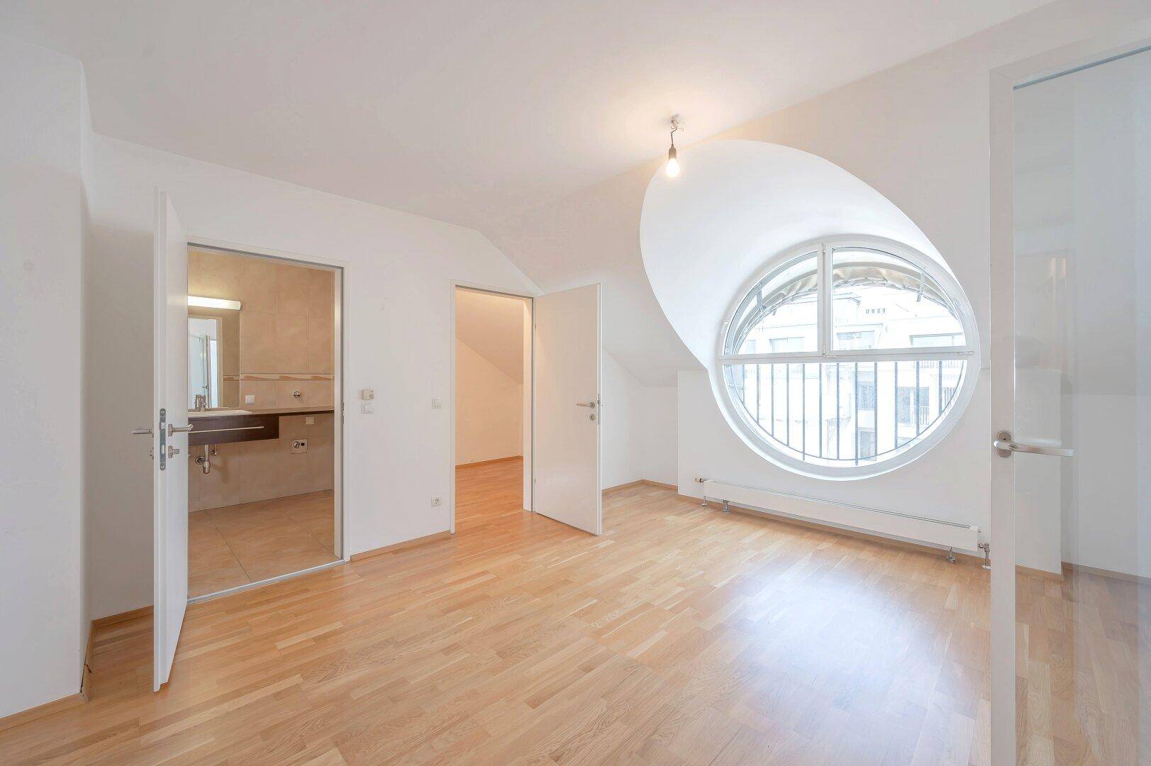 Schlafzimmer mit Parkettboden, markantem Rundfenster und direktem Zugang zum Badezimmer.