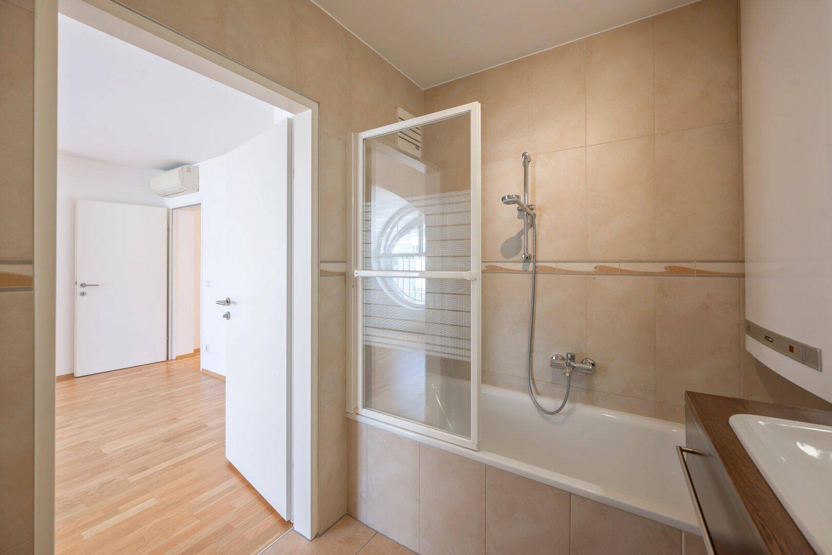Badezimmer mit Badewanne, Duschabtrennung und Blick durch die Tür in den angrenzenden Raum.
