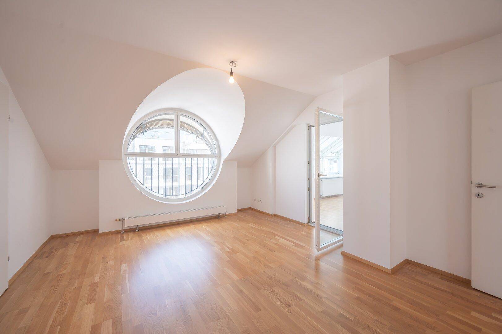 Geräumiges Schlafzimmer mit Parkettboden und einem auffälligen runden Fenster.