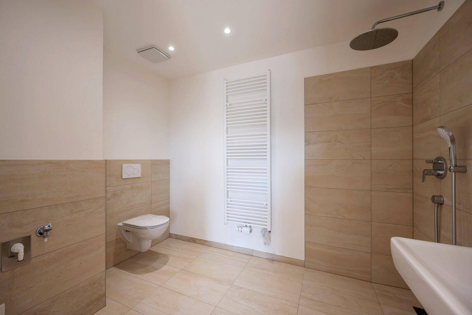 Geräumiges Badezimmer mit Wand-WC, ebenerdiger Dusche, Waschbecken und Handtuchheizkörper.