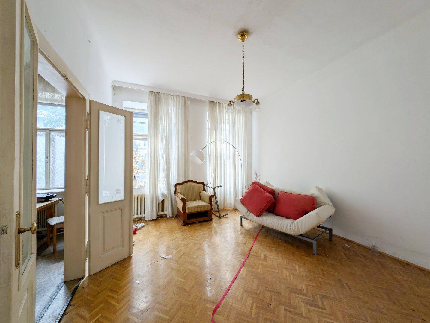 Helles Zimmer mit Fischgrätparkett, hohen Decken und einfacher Möblierung, ideal für Renovierung.