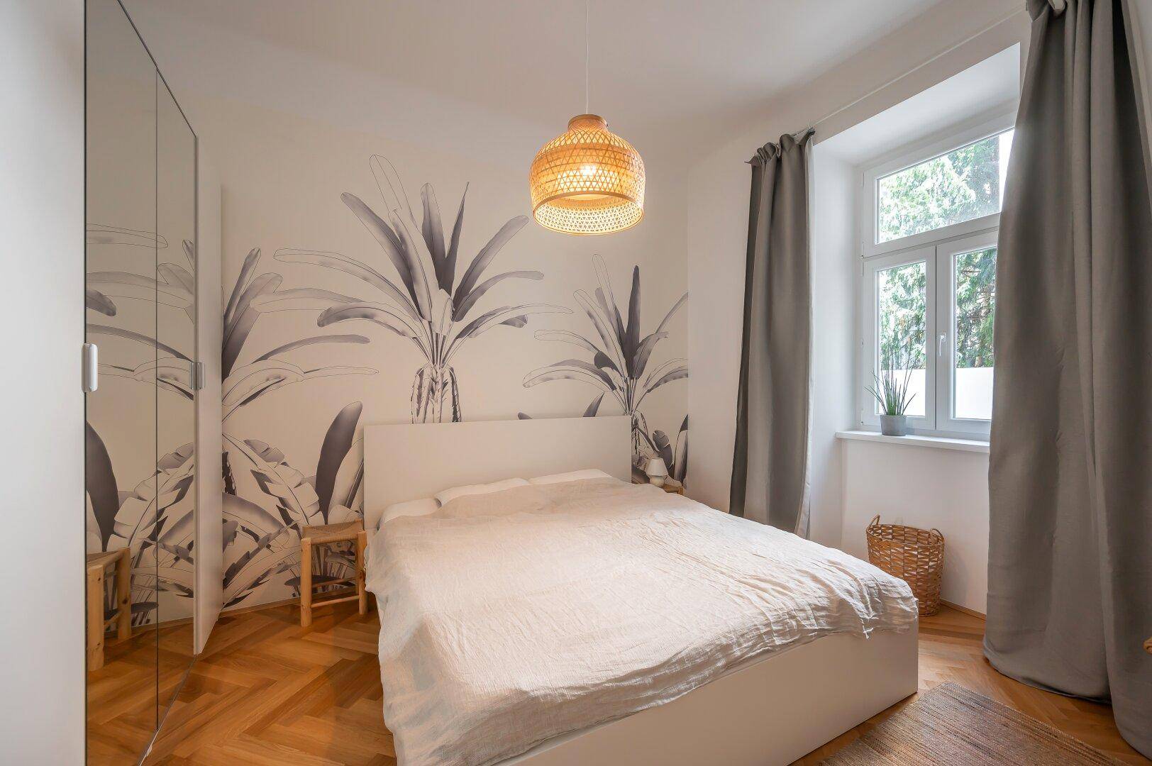 Helles Schlafzimmer mit Doppelbett, Spiegelschrank und dekorativer Wandtapete mit Pflanzenmotiv.