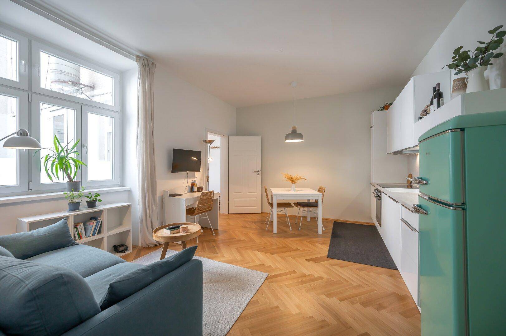 Offener Wohnbereich mit integrierter Küche, Essplatz und Sofa auf hellem Parkettboden.