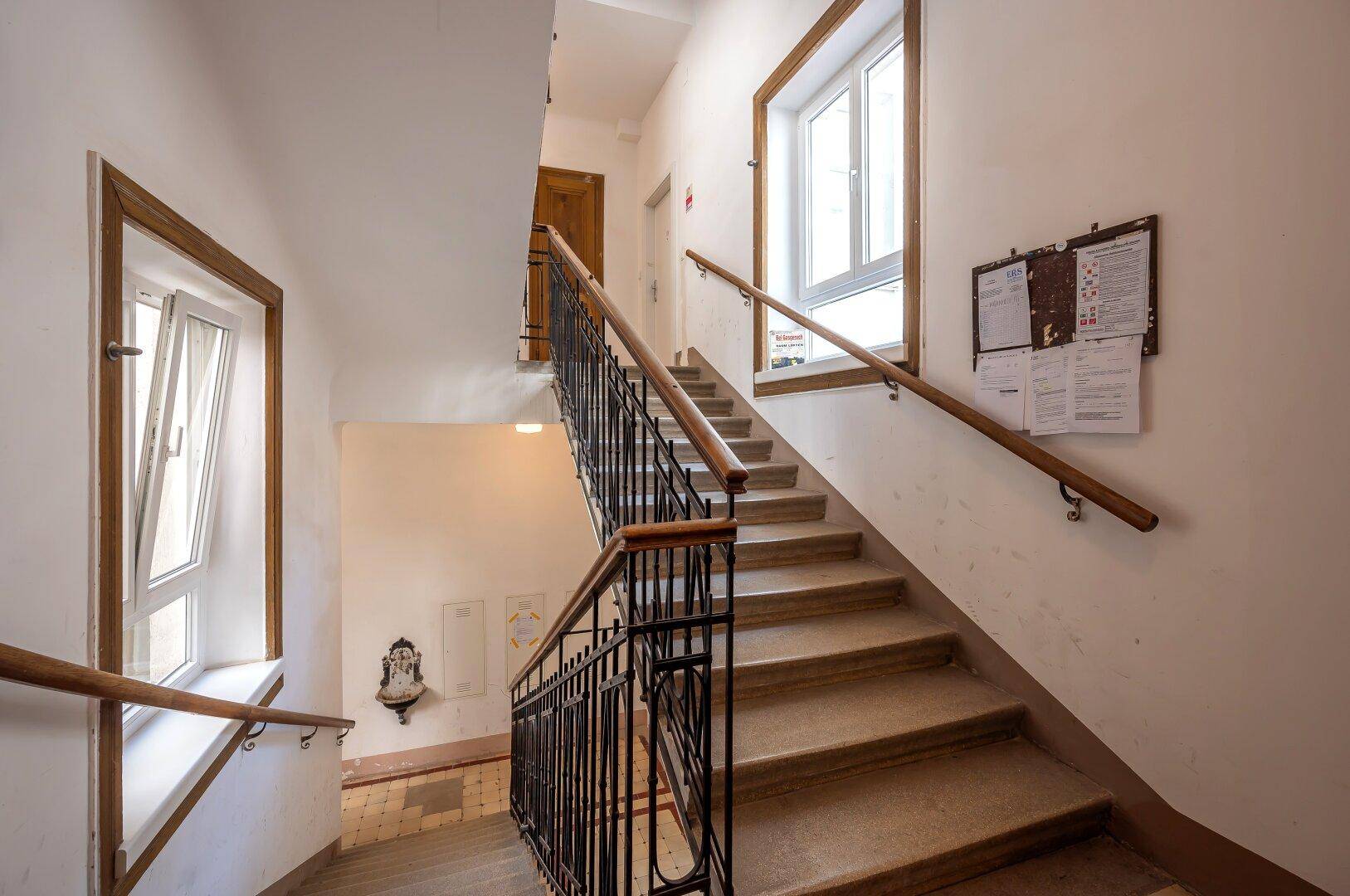Helles Treppenhaus mit Holzgeländer und großen Fenstern, typisch für ein Altbaugebäude.