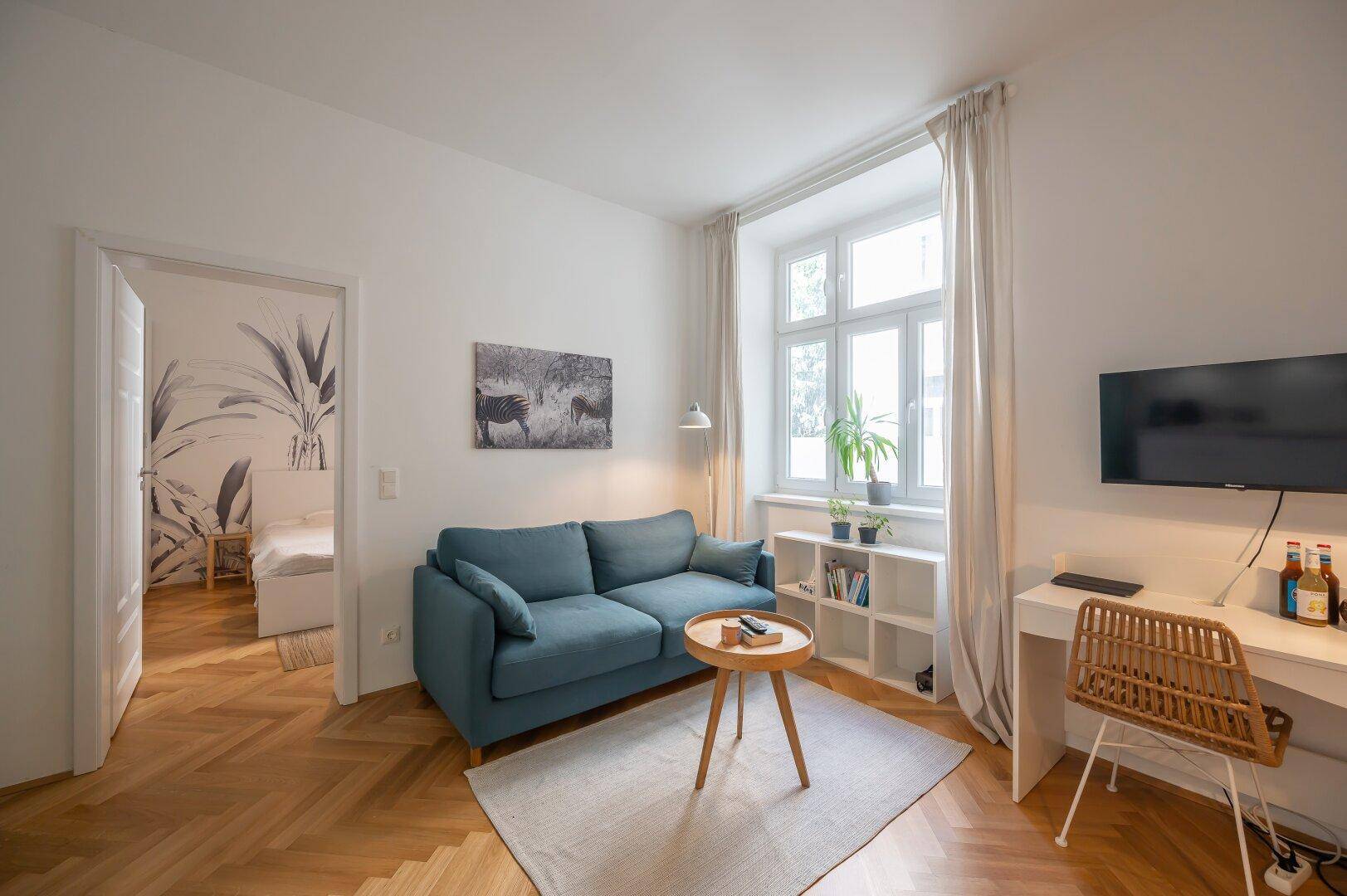 Gemütliches Wohnzimmer mit blauem Sofa, Schreibtisch und Zugang zu einem angrenzenden Schlafzimmer.