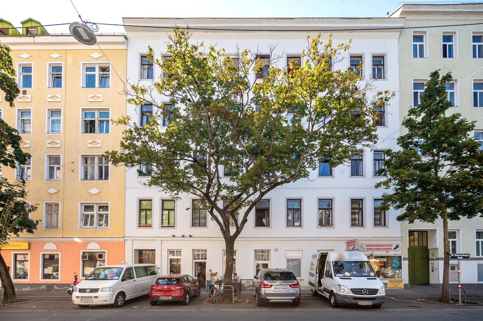 Fassade eines Wohngebäudes an einer Straße mit parkenden Autos und einem großen Baum.