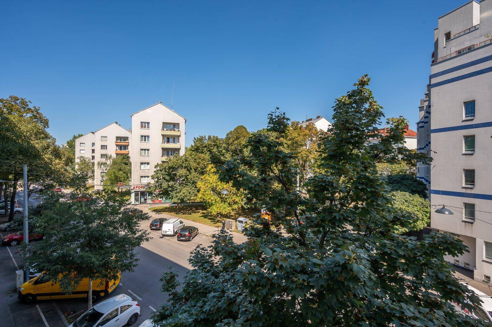 Blick auf eine belebte Straße mit parkenden Autos und Bäumen, umgeben von Wohngebäuden.