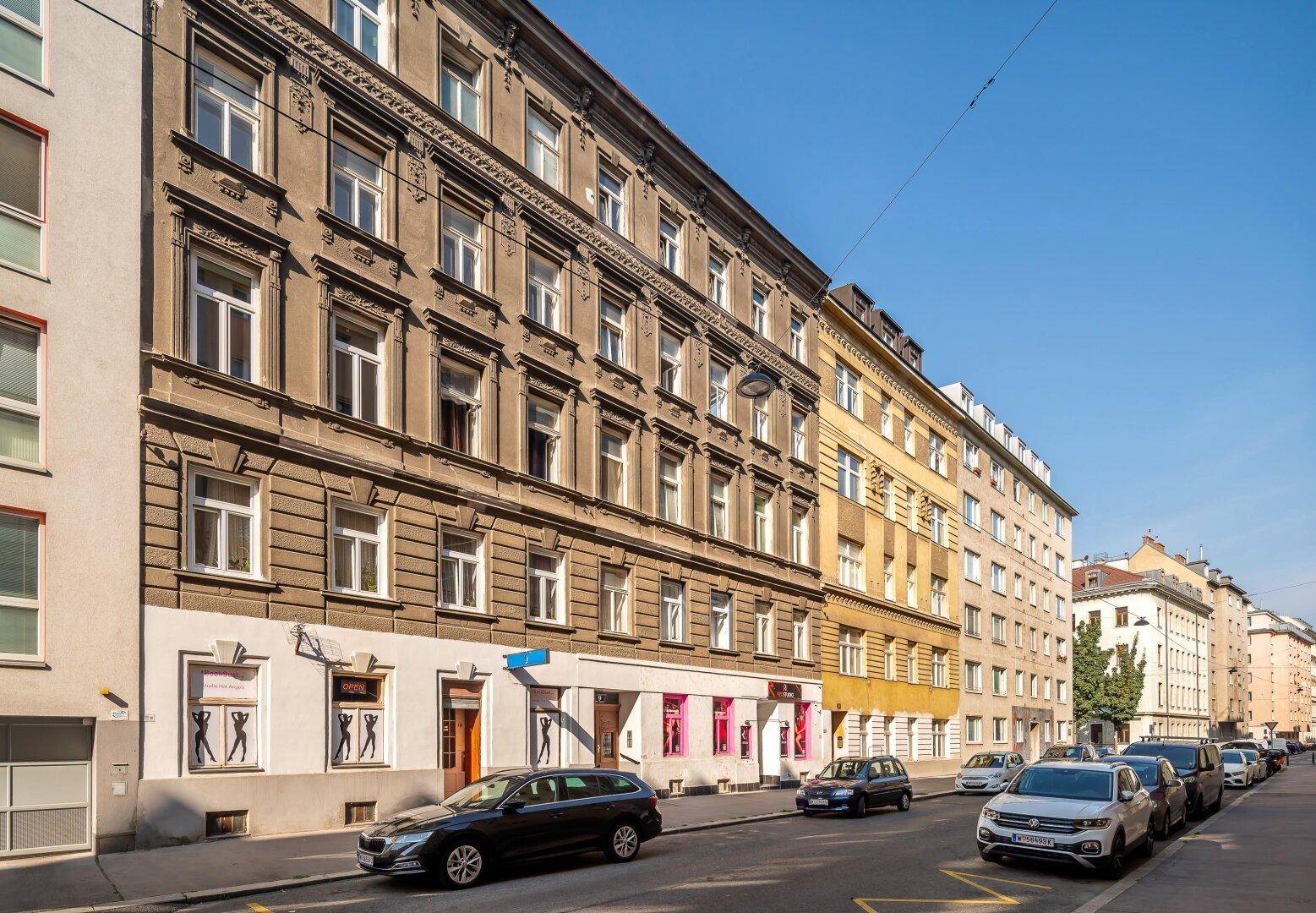 Historische Straßenansicht mit einem Altbau, Geschäften im Erdgeschoss und parkenden Fahrzeugen.