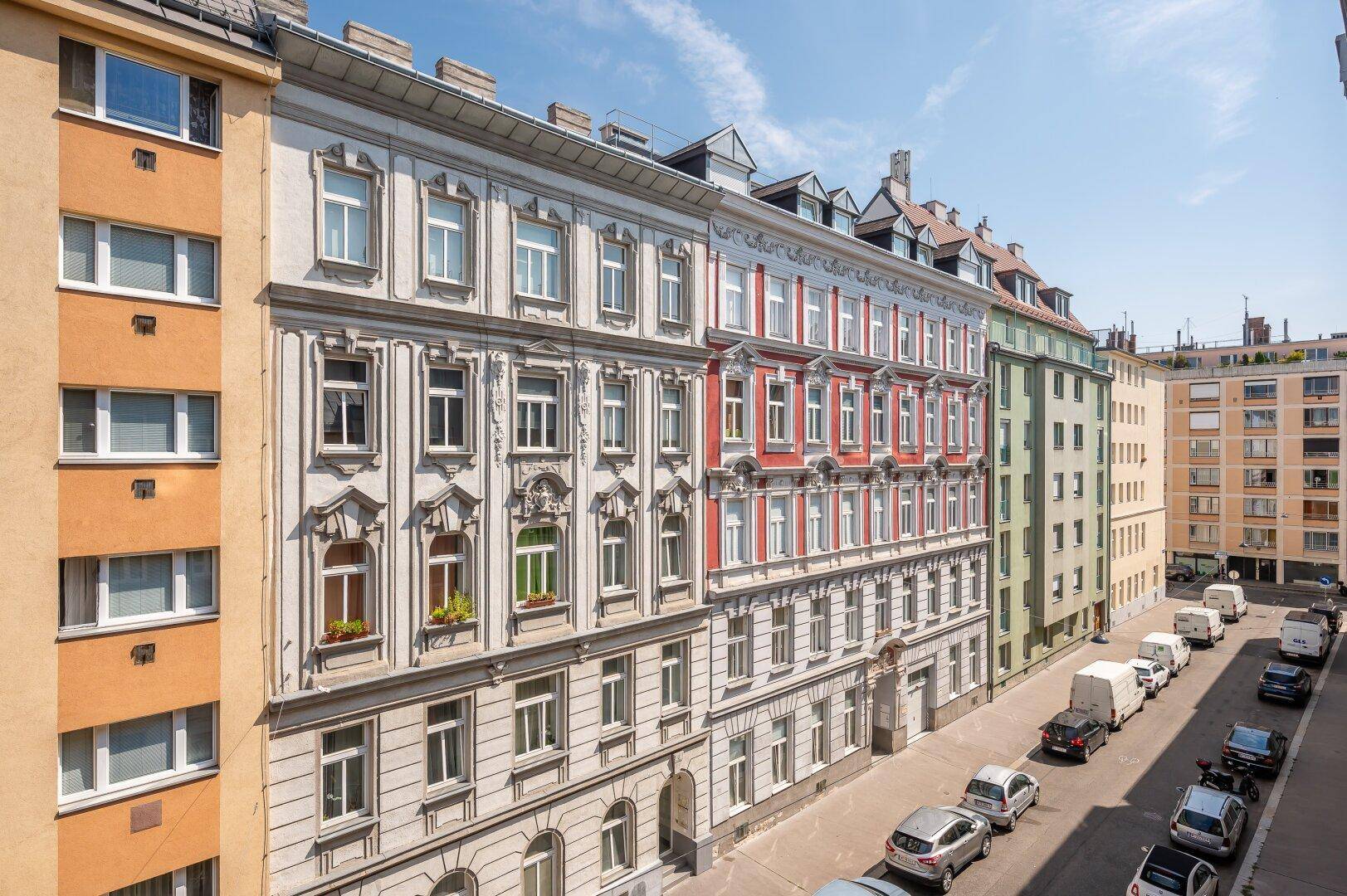 Historische Wohngebäude mit verzierten Fassaden entlang einer belebten Straße mit parkenden Autos.