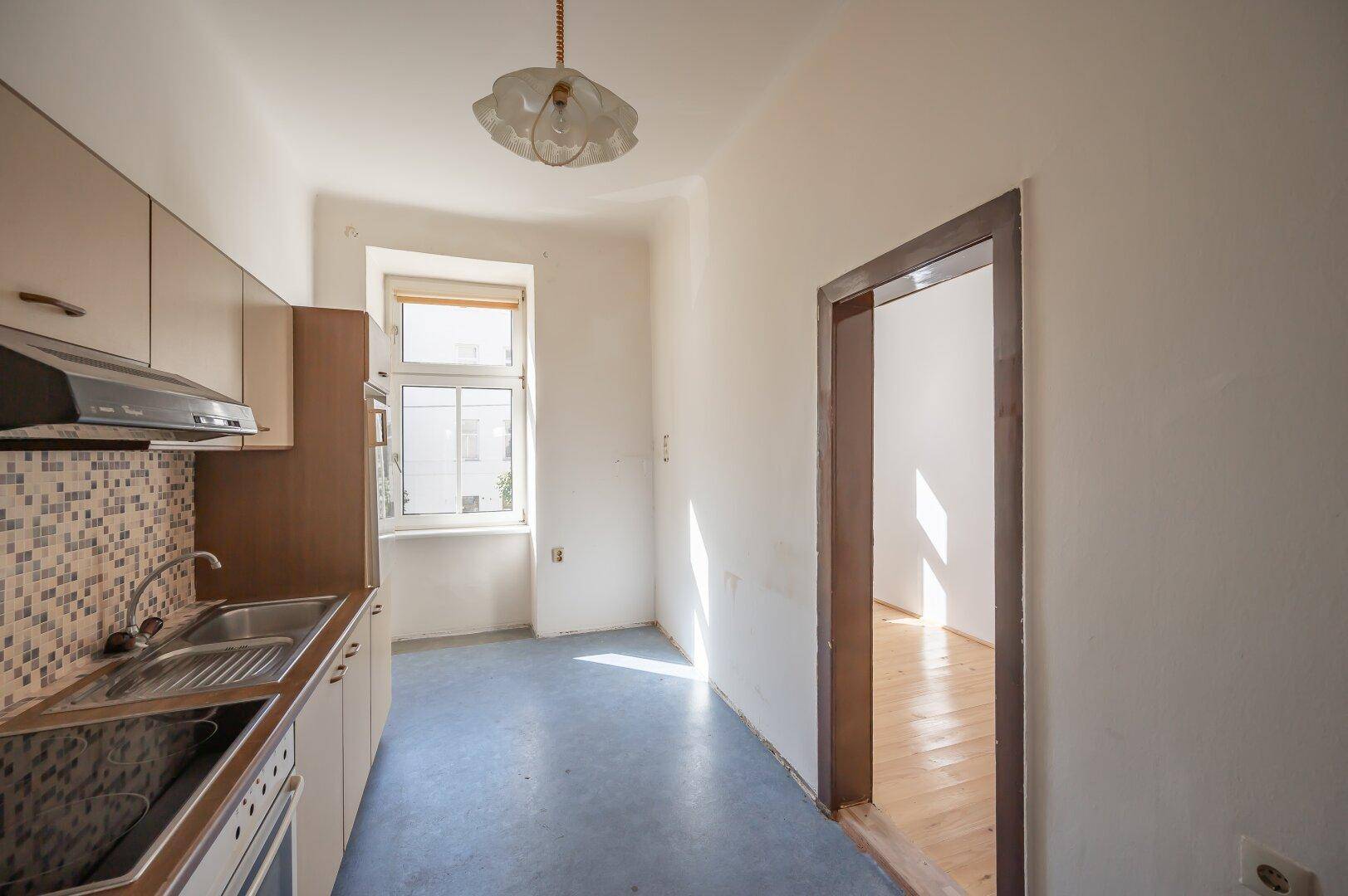 Kleine Küche mit Fenster, älteren Einbaumöbeln und blauer Bodenfliese, die renoviert werden muss.