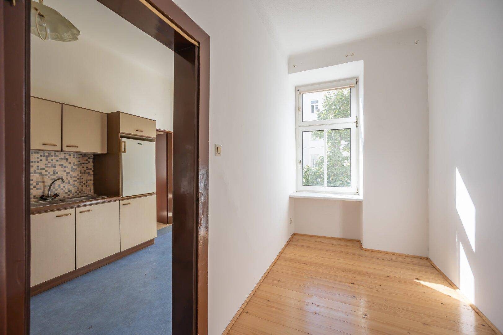 Durchgang zu einer Küche und einem leeren Raum mit Holzboden und Fenster.