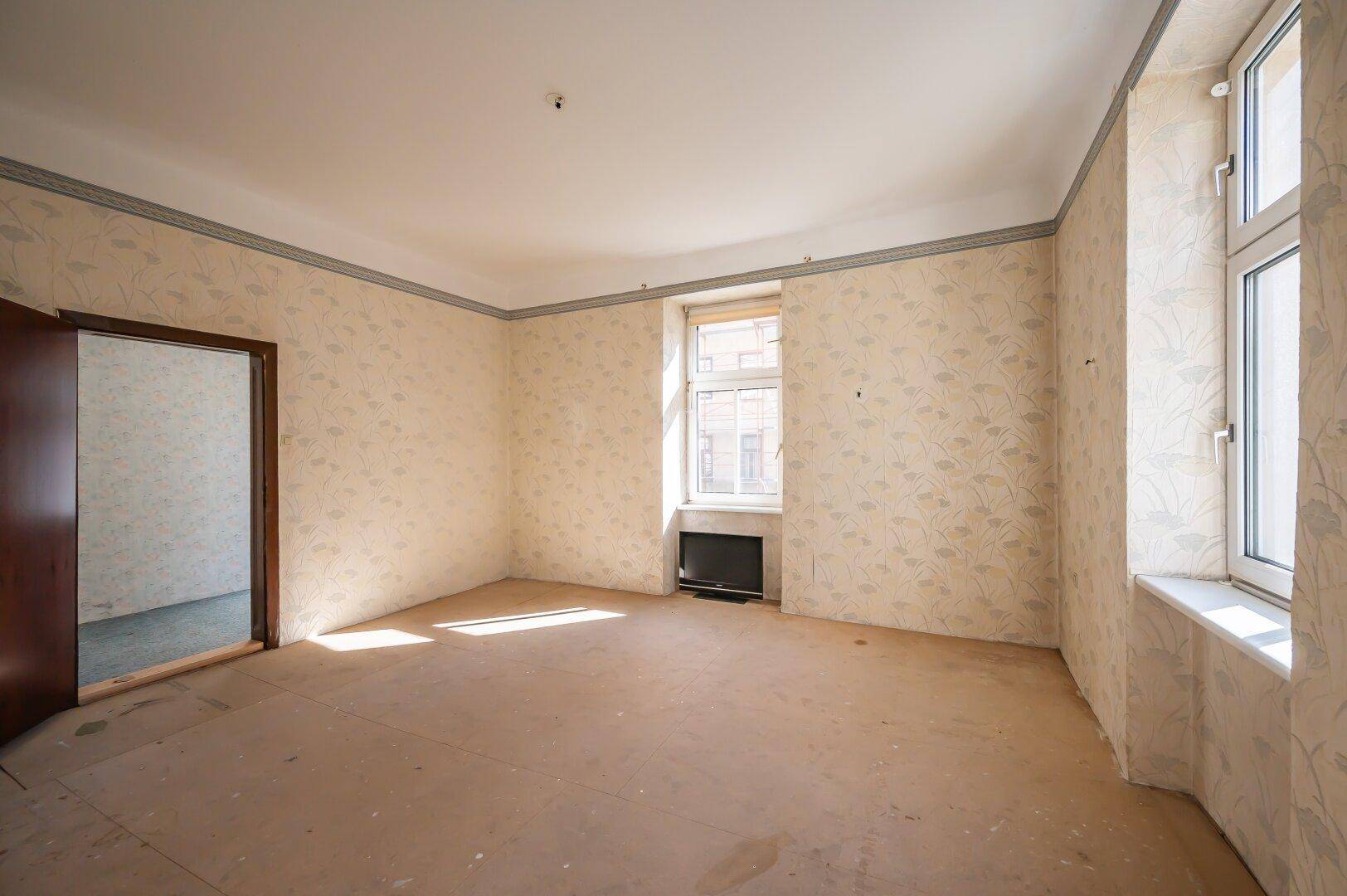 Leeres Zimmer mit alter Tapete, hohen Decken und zwei Fenstern, bereit für eine Renovierung.