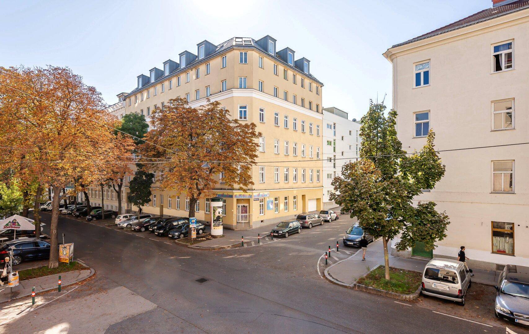 Eckgebäude in einer städtischen Umgebung mit parkenden Autos und Bäumen im Herbstlaub.