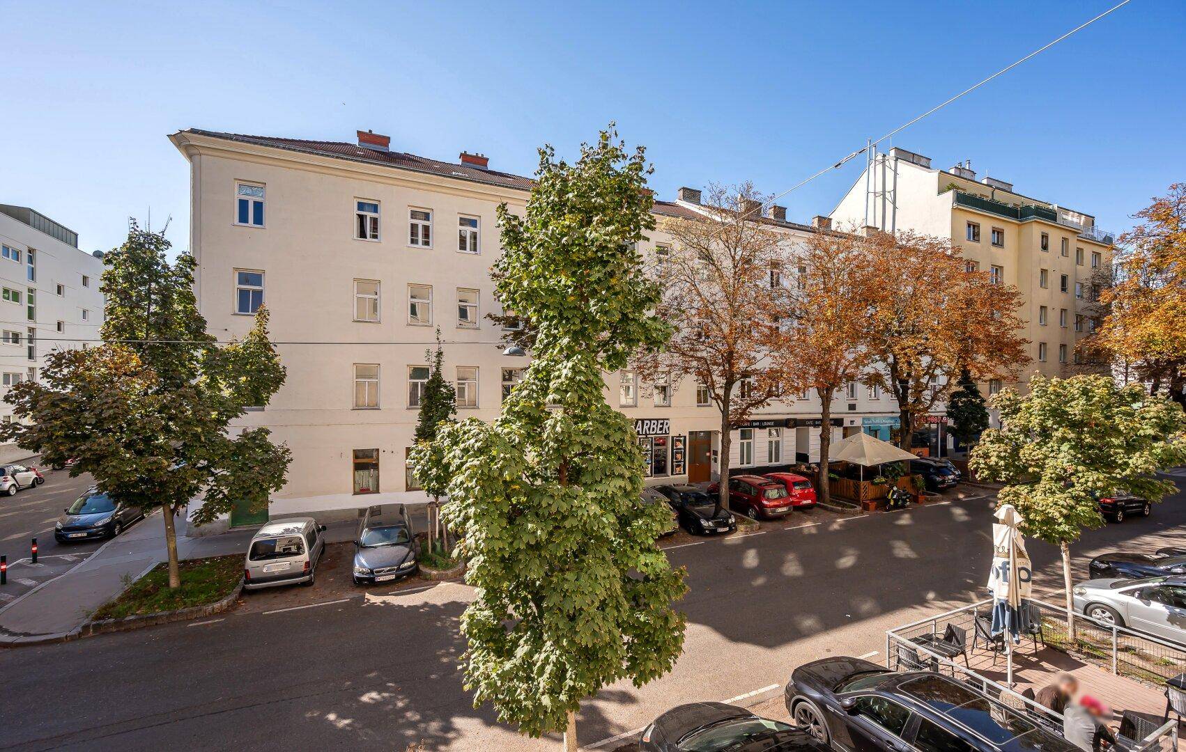 Wohngebäude an einer Straße mit parkenden Autos und Bäumen im Herbstlaub.