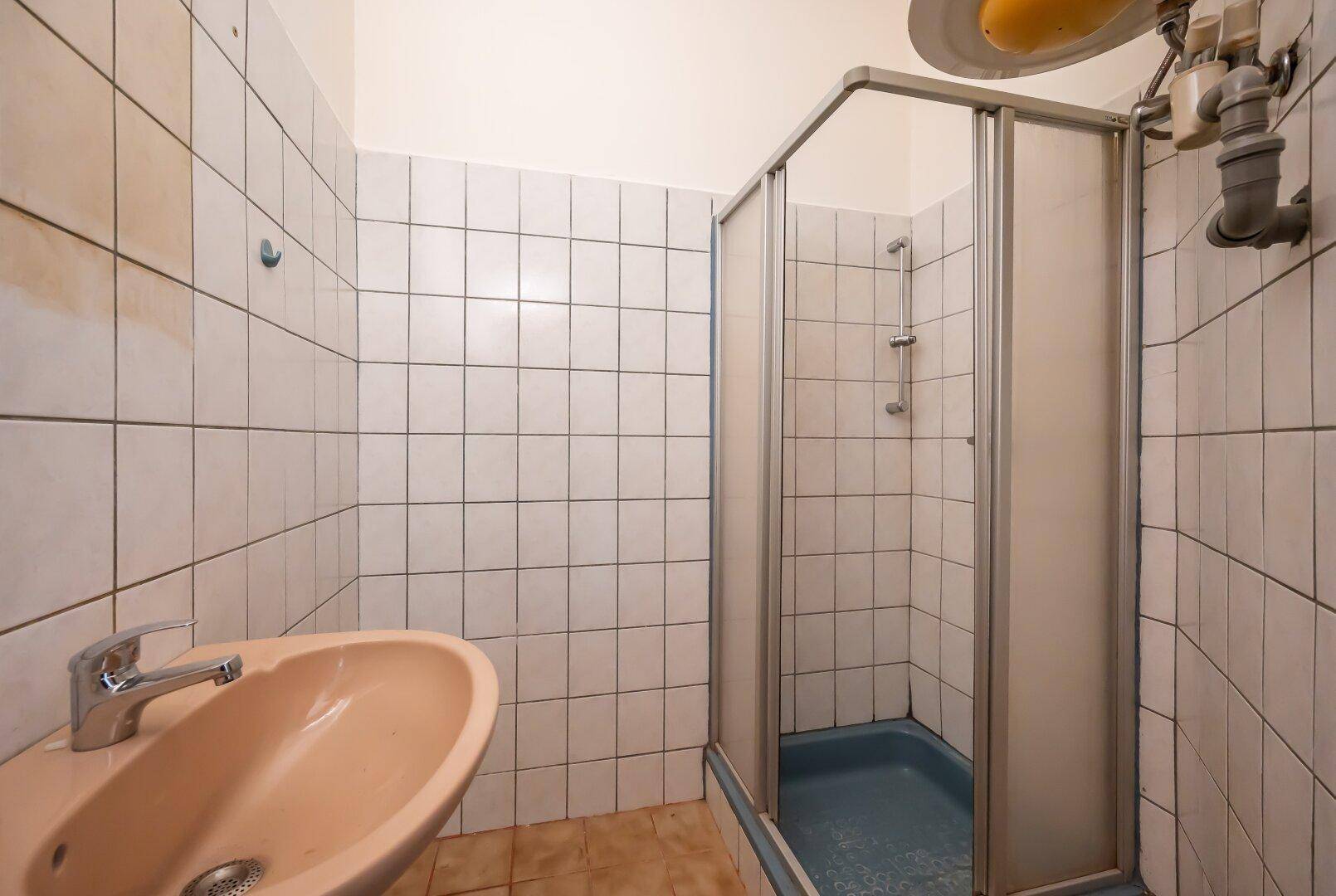 Einfaches Badezimmer mit Duschkabine, Waschbecken und weißen Fliesen, das renoviert werden muss.