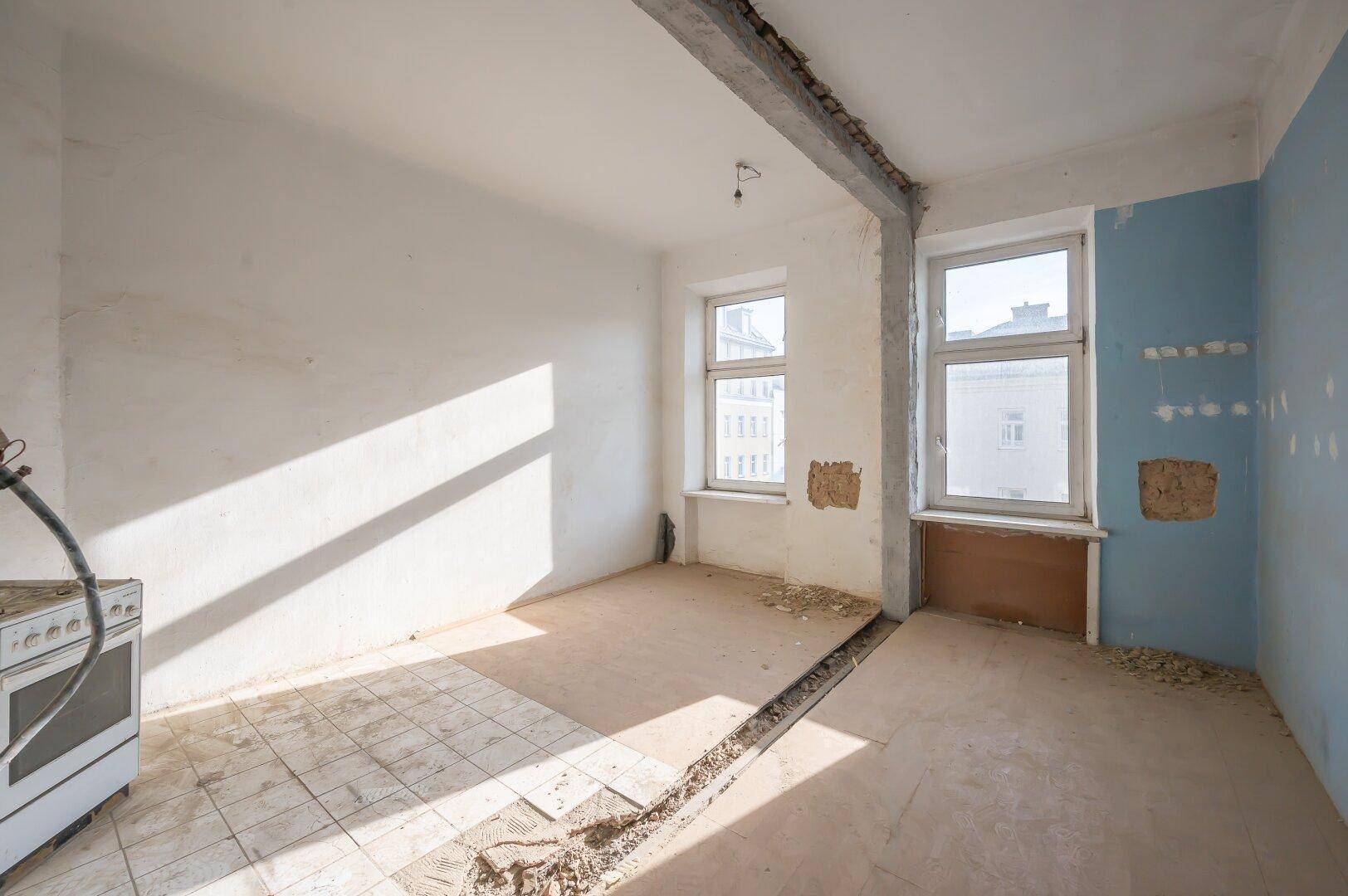 Renovierungsbedürftige Küche mit zwei Fenstern, freigelegtem Träger und teilweise gefliestem Boden.
