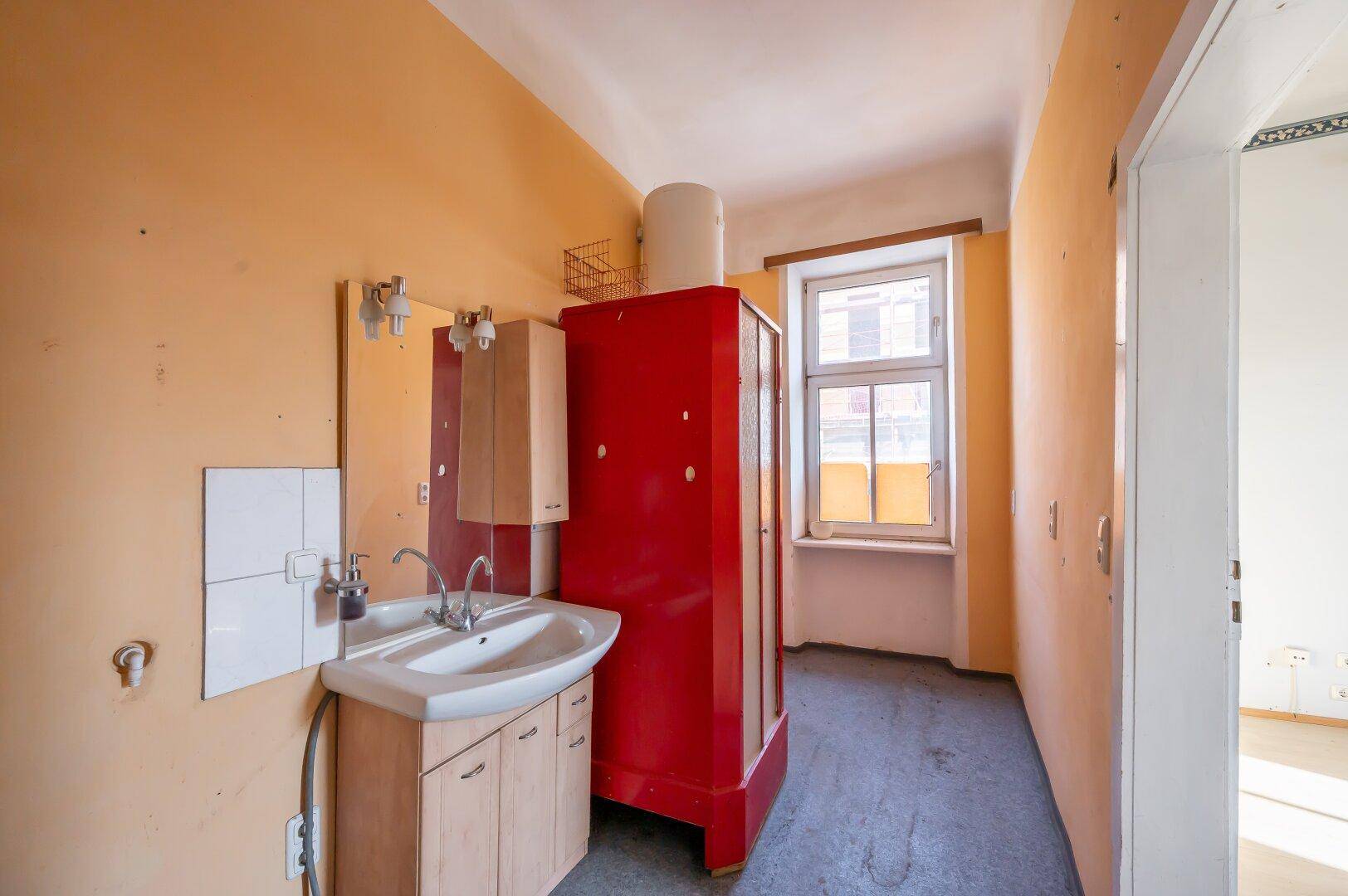 Badezimmer mit Waschbecken, Spiegel, rotem Schrank und einem Fenster für Tageslicht.