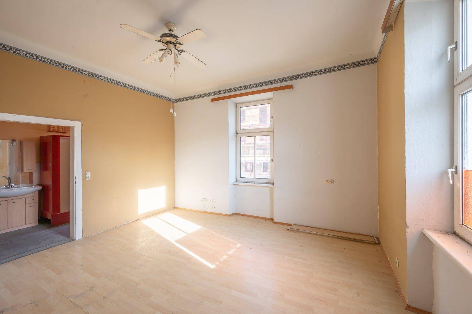Geräumiges Zimmer mit hellem Holzboden, Deckenventilator und Zugang zu einem Nebenraum.