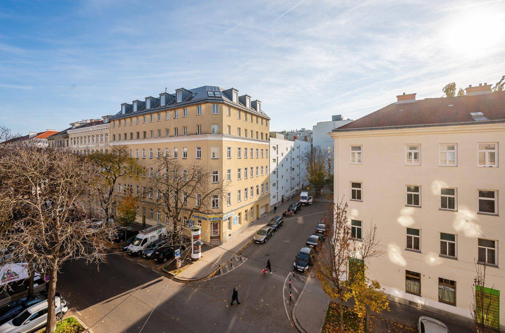 Panoramablick auf eine städtische Kreuzung mit Gebäuden, Bäumen und parkenden Fahrzeugen.
