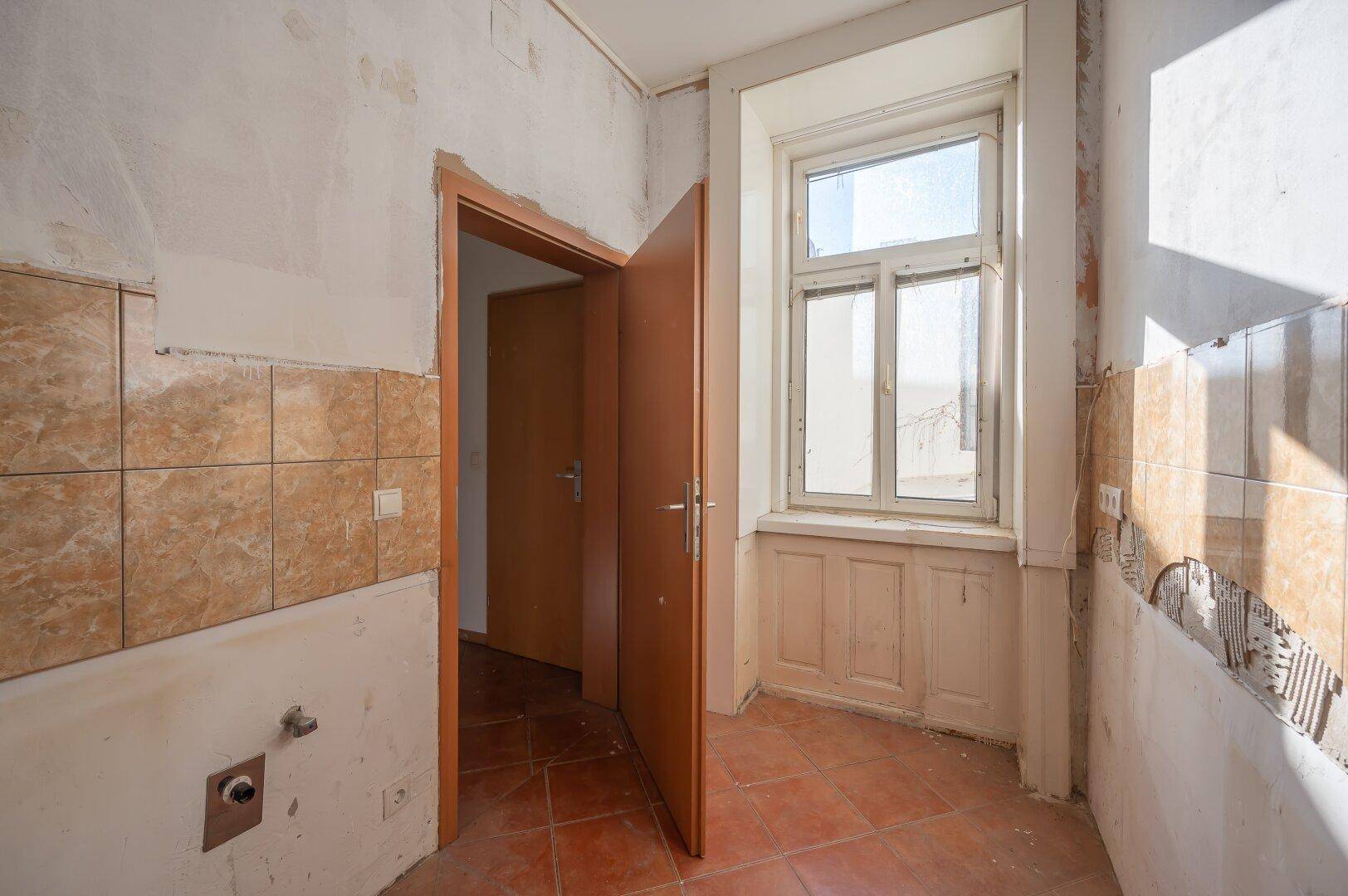 Küche in Renovierung mit Fenster, alten Fliesen und freigelegten Wänden, viel Tageslicht.