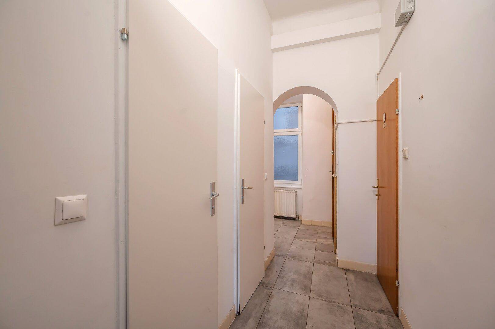 Langer Flur mit Fliesenboden, mehreren Türen und einem Bogen, der zu einem Fenster führt.