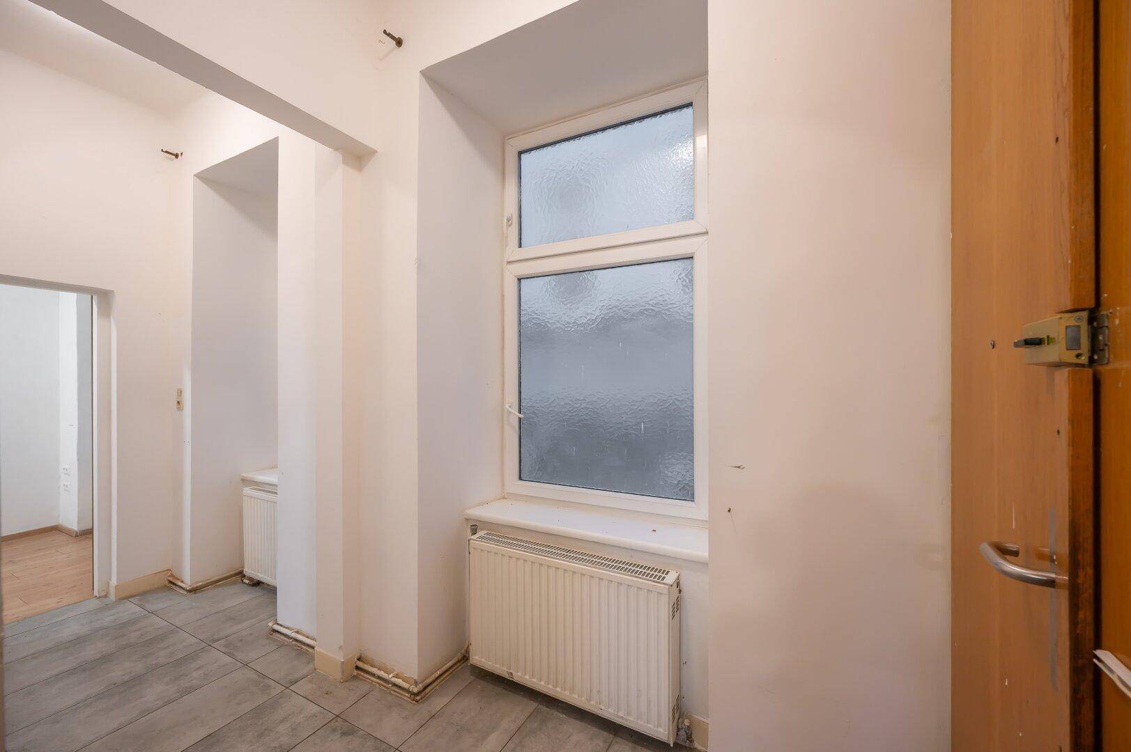 Flur mit Fliesenboden, einem Fenster und einem Heizkörper, der für natürliche Belichtung sorgt.