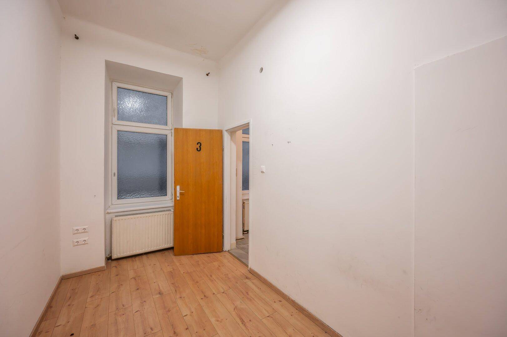 Leeres Zimmer mit Holzboden, einem Fenster und einer Holztür, bereit zur Nutzung.