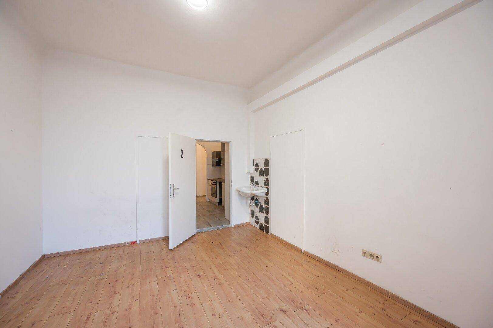 Leeres Zimmer mit Holzboden und Blick durch eine offene Tür in einen gefliesten Bereich.