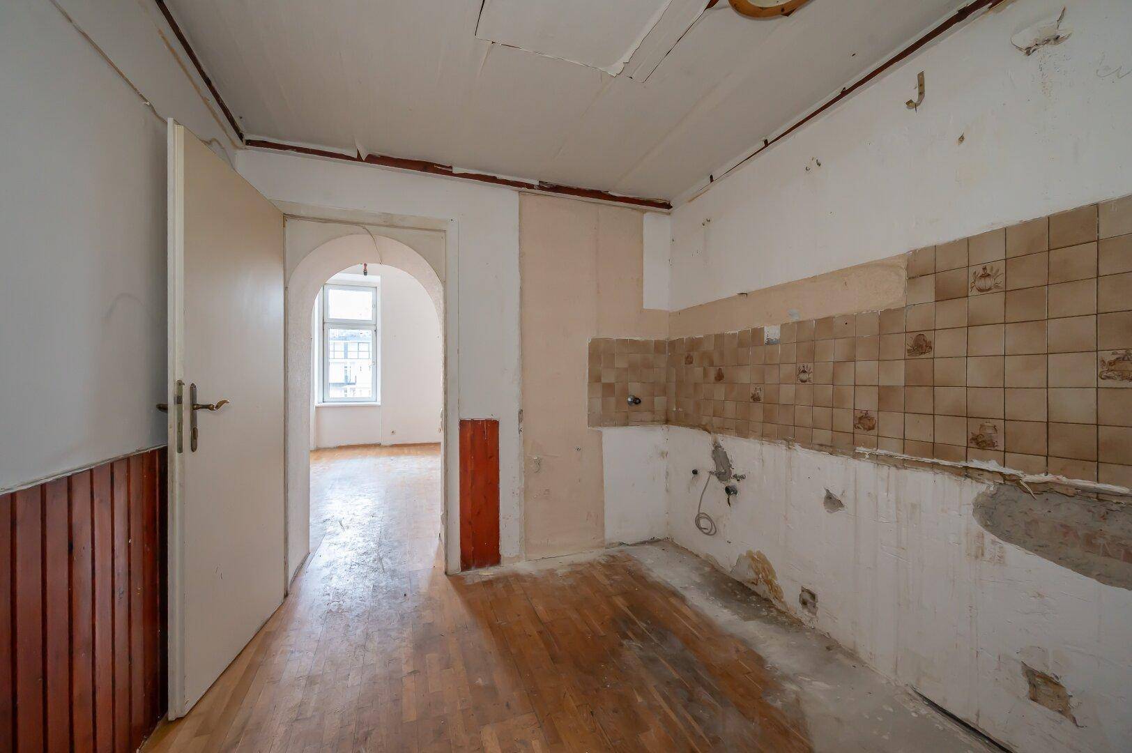 Leere Küche mit alten Wandfliesen und Holzboden, die auf eine Modernisierung wartet.