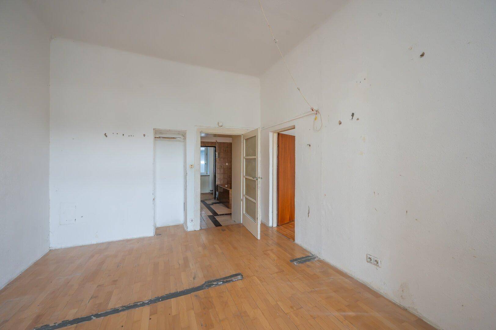 Leeres Zimmer mit Holzboden und mehreren Türen, das auf eine umfassende Renovierung wartet.