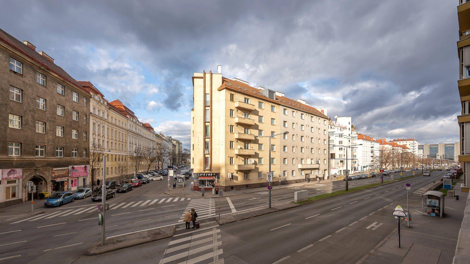 Weitläufige Straßenansicht mit Gebäuden, Verkehr und bewölktem Himmel in einer städtischen Umgebung.