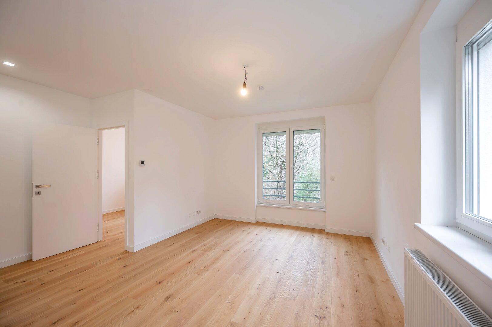 Leerer Raum mit hellem Holzboden, weißen Wänden und großem Fenster.