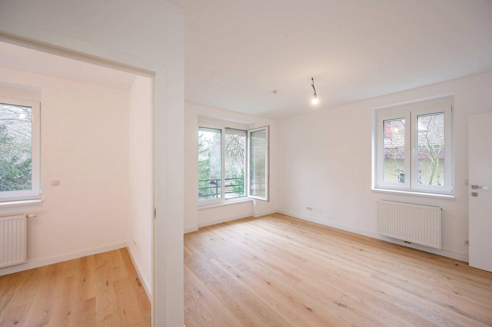 Heller, leerer Raum mit Holzboden, weißen Wänden und Fenstern mit Blick ins Grüne.