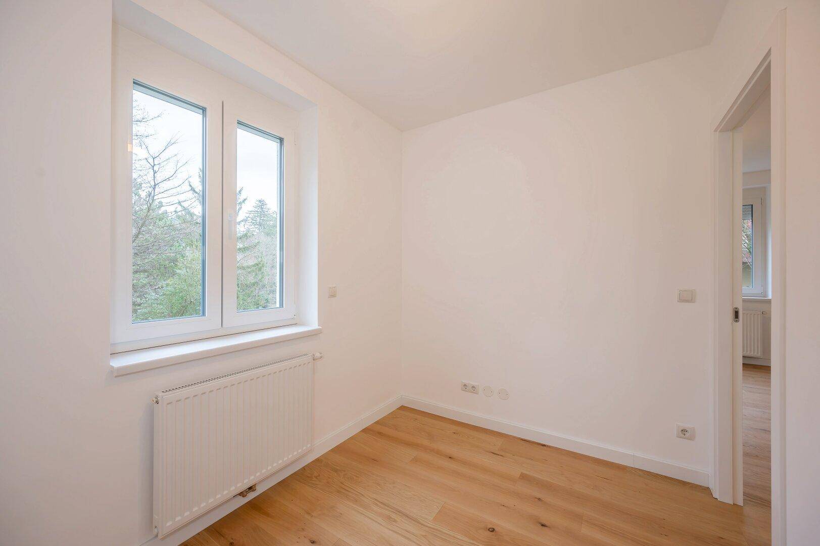 Heller, leerer Raum mit Holzboden, weißem Heizkörper und Fenster mit Blick ins Grüne.