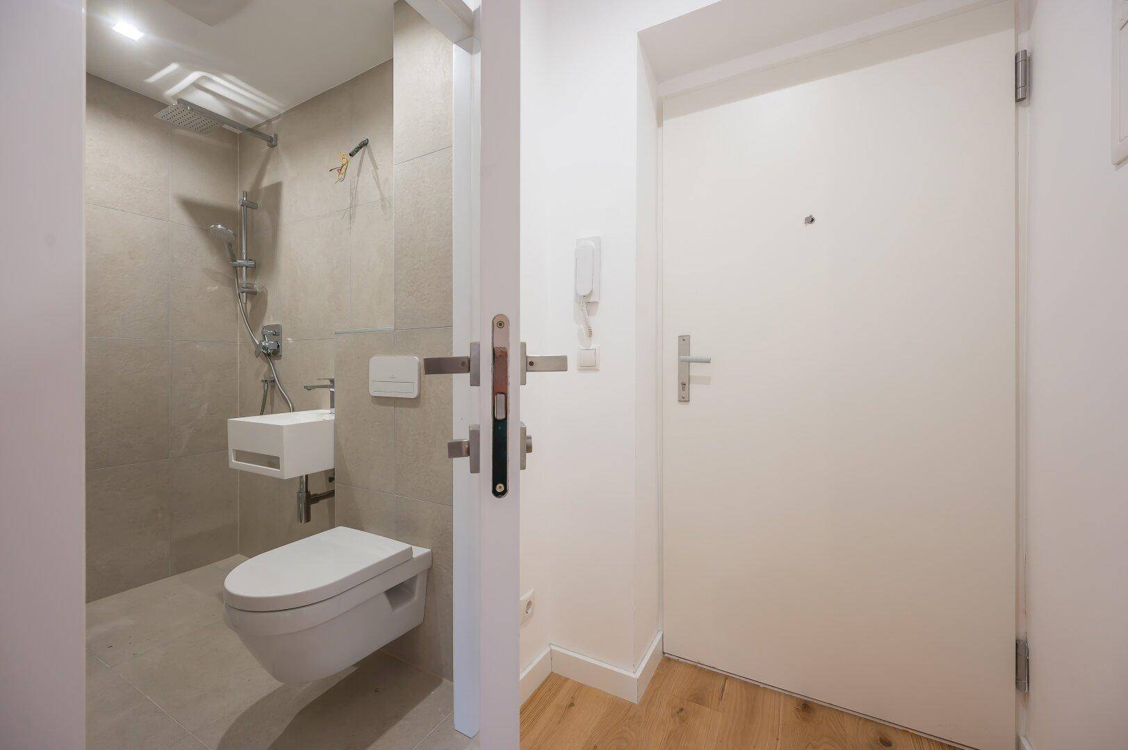 Modernes Badezimmer mit begehbarer Dusche, Hänge-WC und kleinem Waschbecken, direkt neben der Eingangstür.