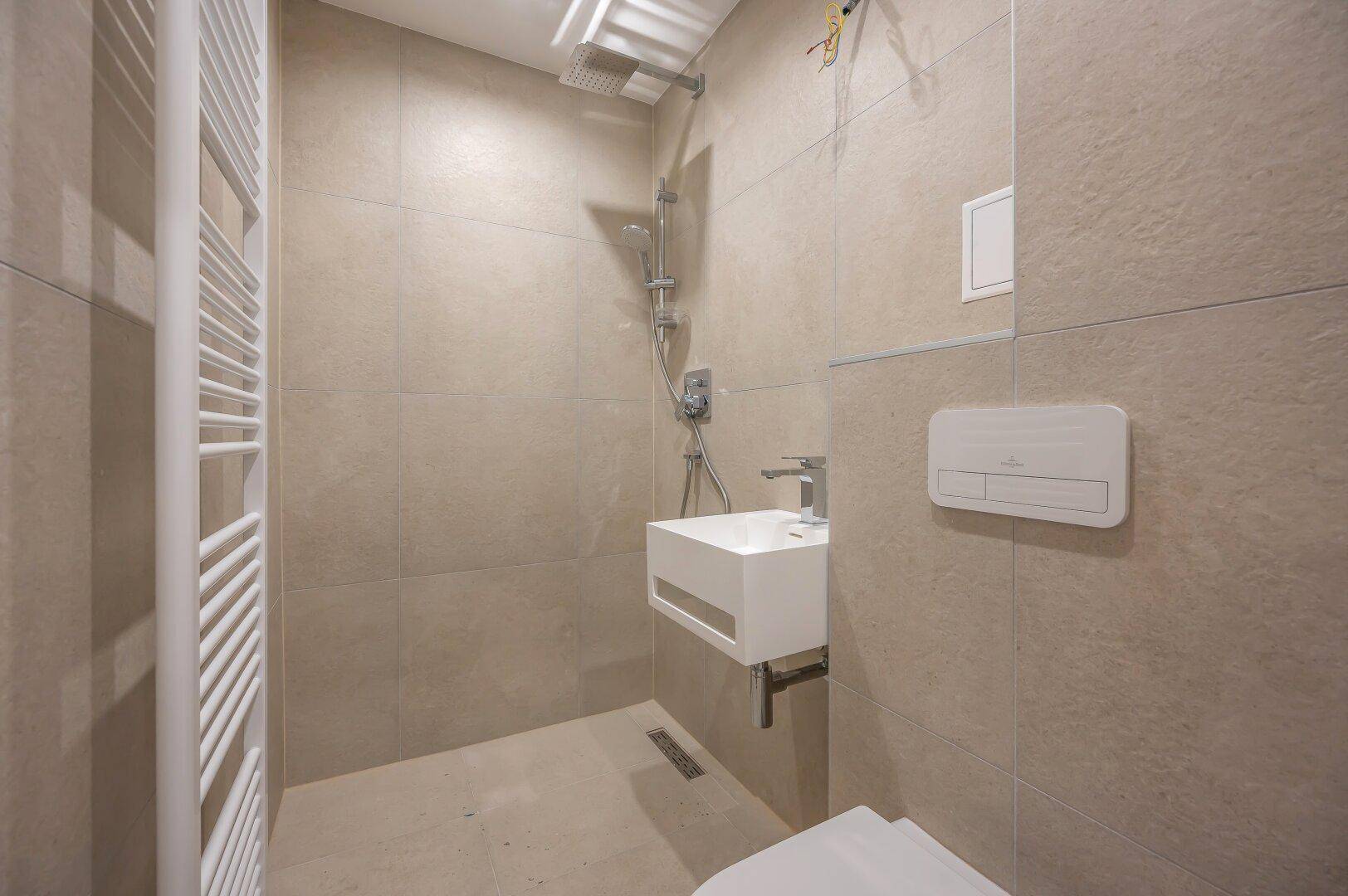 Detaillierte Ansicht des modernen Badezimmers mit Regendusche, Handtuchheizkörper und grauen Fliesen.