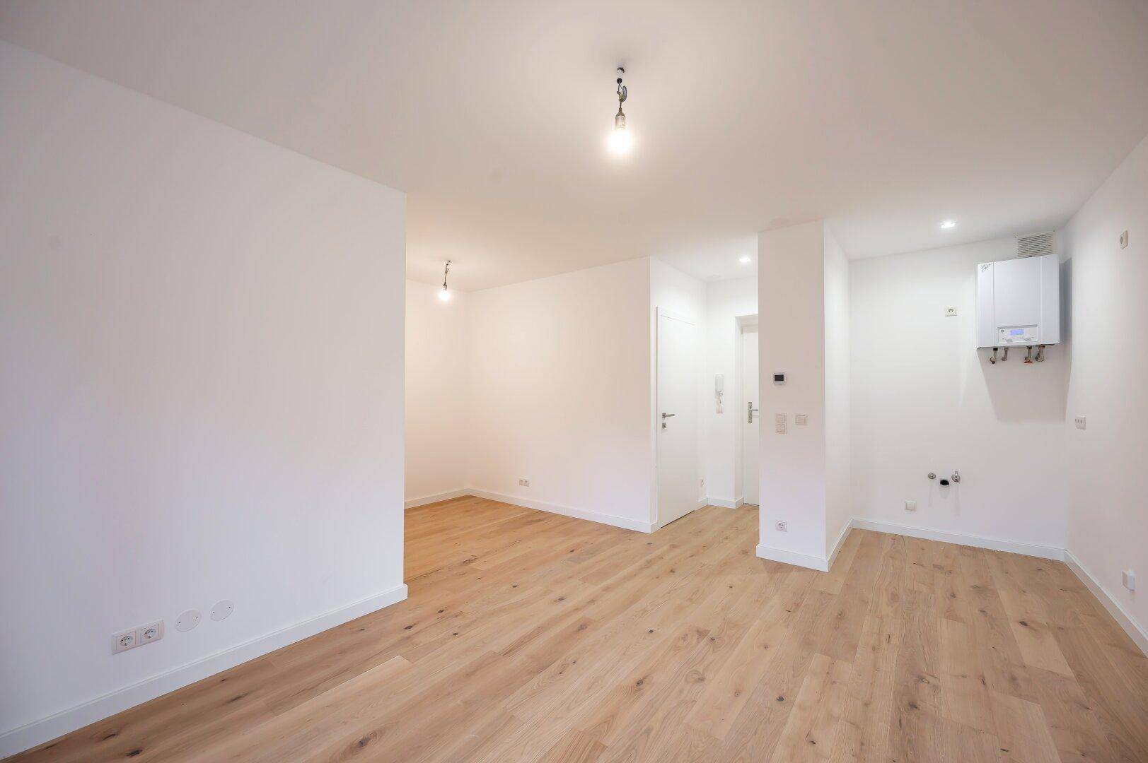 Unmöblierter Raum mit Holzfußboden, Heizungsanlage und vorbereiteten Anschlüssen für Haushaltsgeräte.
